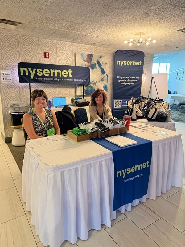 Nysernet Project Manager Elyse Miller and Administrative Assistant Mindy Cooper sit at a registration table decorated in Nysernet signage.