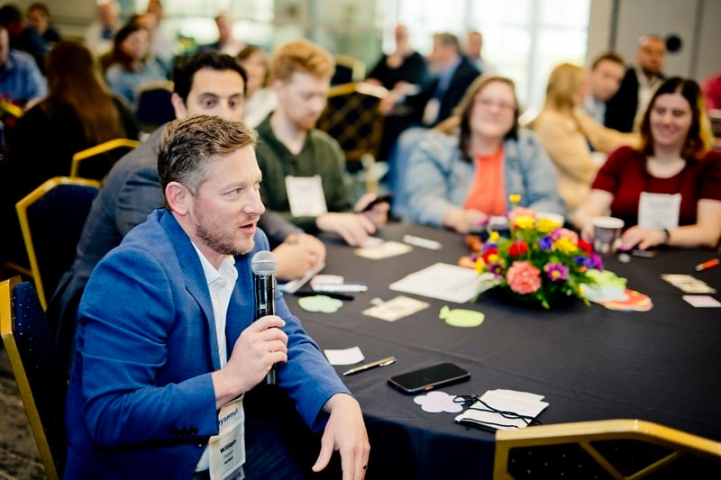 Nysernet members gathered around a banquet table. In the foreground, one of the members speaks into a microphone.