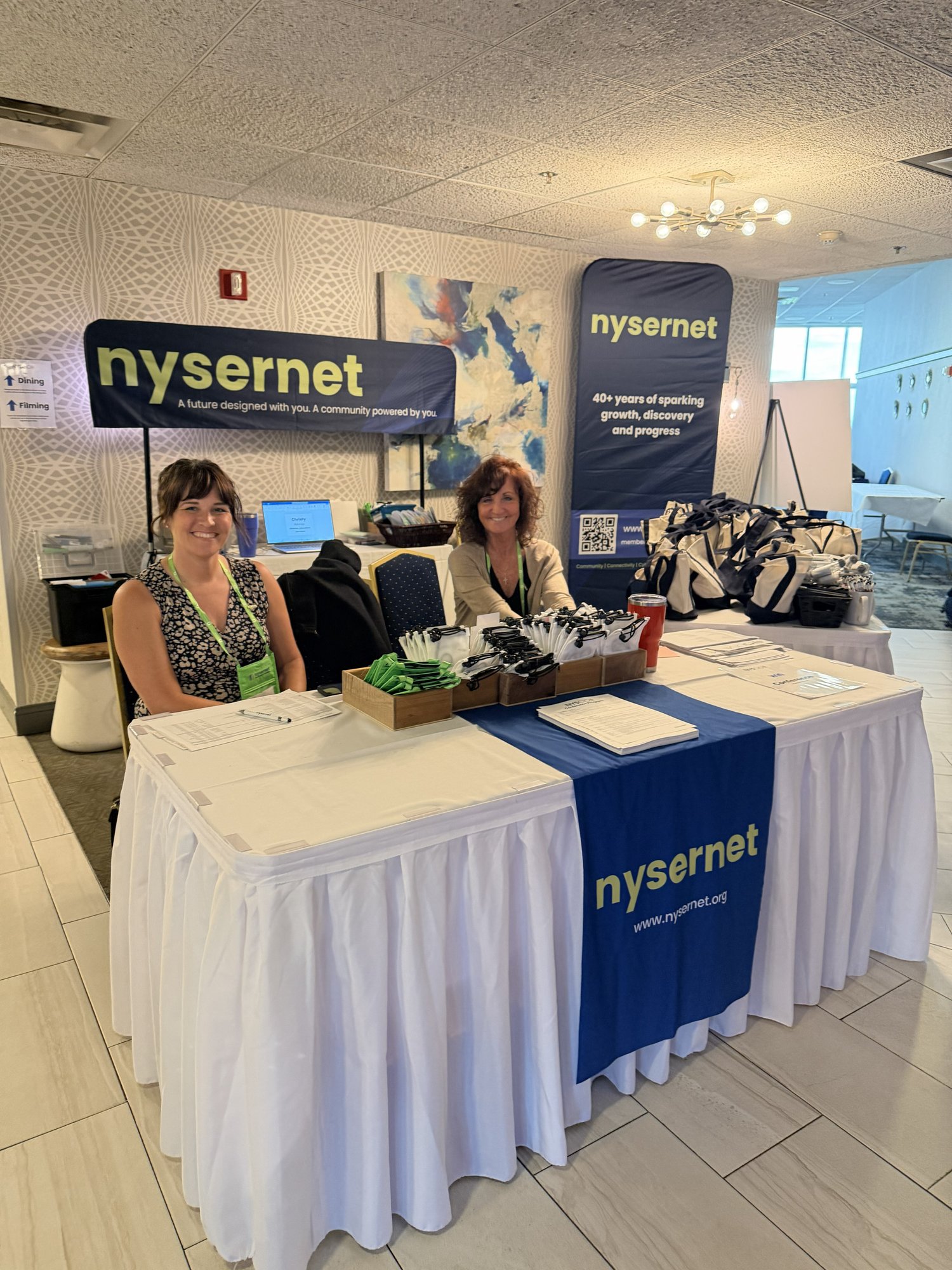 Nysernet Project Manager Elyse Miller and Administrative Assistant Mindy Cooper sit at a registration table decorated in Nysernet signage.